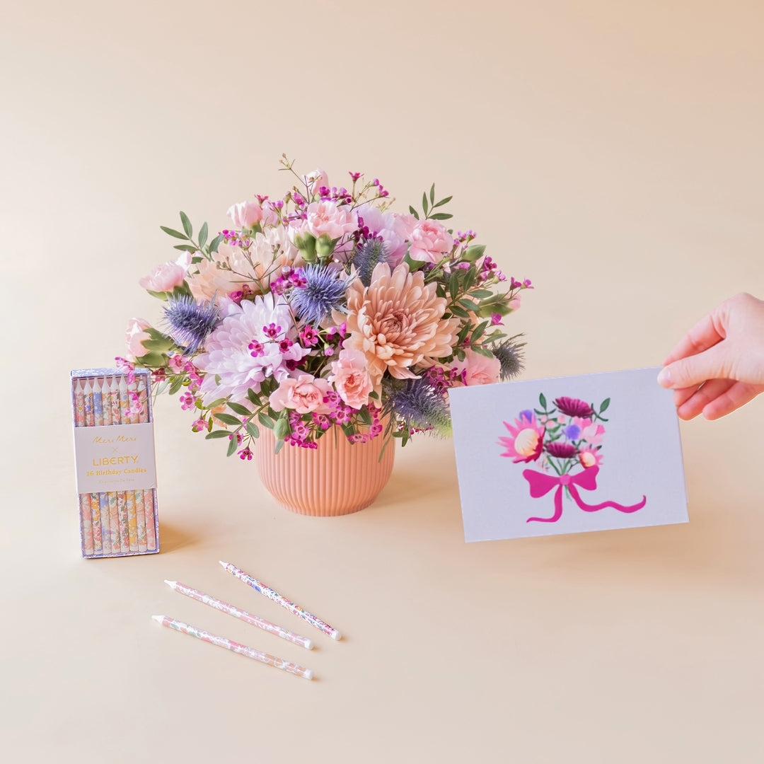 Arreglo de flores en tonos rosas y malvas Guiño en base estriado rosa, junto a una caja de velas de cumpleaños florales de Meri Meri x Liberty y una mano sosteniendo la tarjeta ilustrada con un ramo de flores sobre fondo crema.