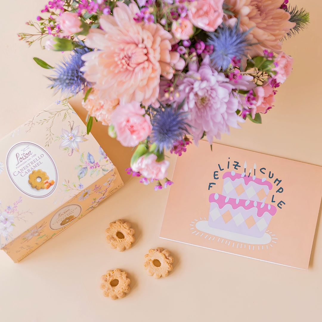 Detalle del arreglo de flores Guiño con crisantemos y cardos azules , junto a una caja de galletas de caramelo Loison y una tarjeta de felicitación de Feliz Cumpleaños sobre un fondo crema.
