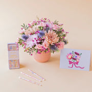 Arreglo de flores en tonos rosas y malvas Guiño en base estriado rosa, junto a una caja de velas de cumpleaños florales de Meri Meri x Liberty y una tarjeta ilustrada con un ramo de flores sobre fondo crema.