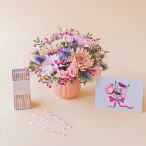 Arreglo de flores en tonos rosas y malvas Guiño en base estriado rosa, junto a una caja de velas de cumpleaños florales de Meri Meri x Liberty y una tarjeta ilustrada con un ramo de flores sobre fondo crema.