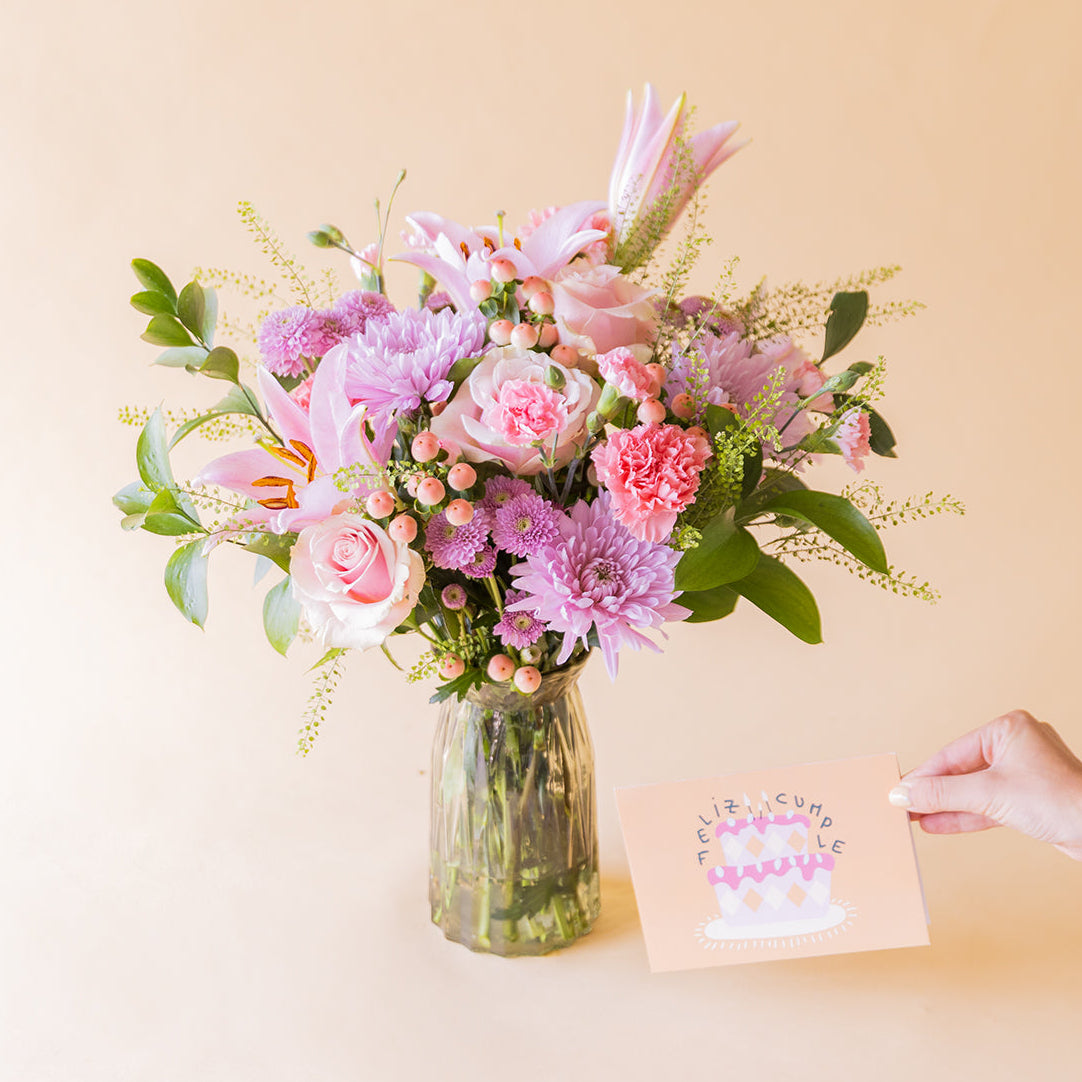 Ramo de flores rosas de cumpleaños de Colvin en jarrón de cristal con lirios, crisantemos lilas, rosas y claveles, junto a tarjeta "Feliz Cumple"
