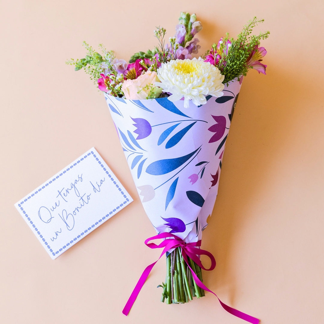 Pack La Costa de Colvin, que incluye un ramo de flores frescas de verano con rosas amarillas, crisantemos blancos y alstroemerias fucsias, envuelto en papel con motivos florales y acompañado de una tarjeta azul con el texto manuscrito "Que tengas un Bonito día" sobre fondo beige.