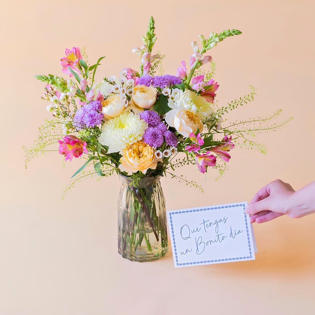 Ramo de flores frescas de Colvin, La Costa con rosas de jardín melocotón, alstroemerias y crisantemos en un jarrón estriado de cristal transparente, una mano sosteniendo una tarjeta de felicitación con la frase "Que tengas un Bonito día" sobre un fondo liso de color crema.