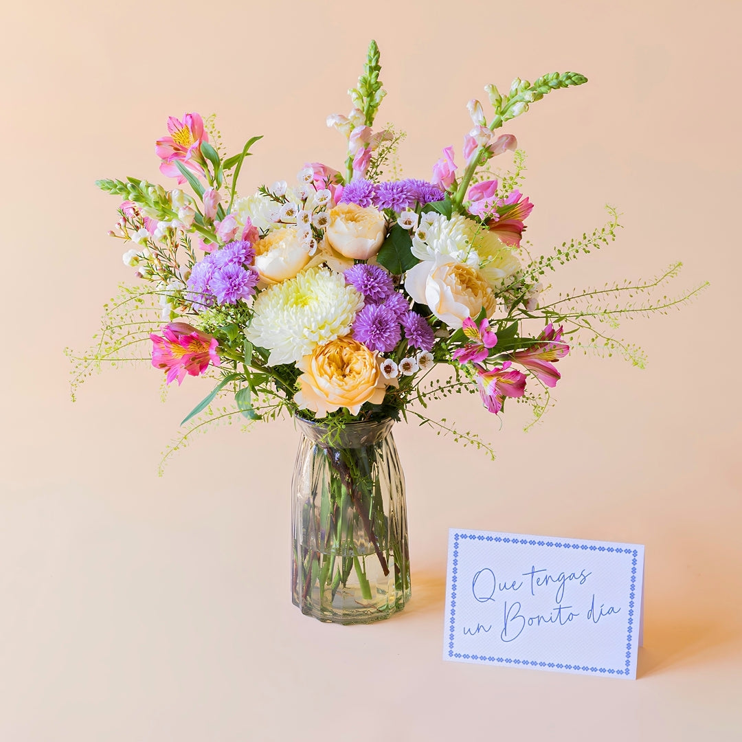 Ramo de flores frescas de Colvin, La Costa con rosas de jardín melocotón, alstroemerias y crisantemos en un jarrón estriado de cristal transparente, junto a una tarjeta de felicitación con la frase "Que tengas un Bonito día" sobre un fondo liso de color crema.