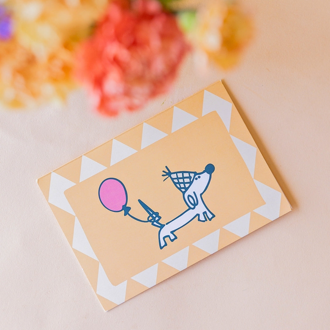 Primer plano de tarjeta ilustrada con perro salchicha con gorro de fiesta y globo rosa, con ramo de claveles naranja y melocotón de Colvin desenfocado al fondo