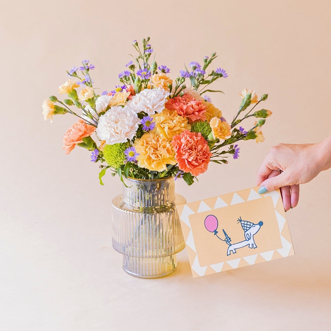 Ramo de claveles naranja, melocotón, blanco y aster lila de Colvin en jarrón acanalado transparente, con mano sosteniendo tarjeta ilustrada de perro salchicha con globo rosa, sobre fondo melocotón
