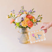 Ramo de claveles naranja, melocotón, blanco y aster lila de Colvin en jarrón acanalado transparente, con mano sosteniendo tarjeta ilustrada de perro salchicha con globo rosa, sobre fondo melocotón