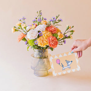 Ramo de claveles naranja, melocotón, blanco y aster lila de Colvin en jarrón acanalado transparente, con mano sosteniendo tarjeta ilustrada de perro salchicha con globo rosa, sobre fondo melocotón