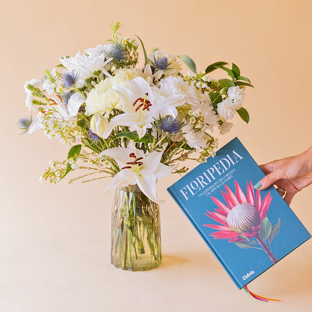 Mano sosteniendo el libro Floripedia de Colvin junto a ramo de flores blancas en jarrón de cristal con lirios, crisantemos y eryngium azul