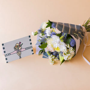 Ramo de flores blancas Colvin con crisantemo, eryngium y wax flower en envoltorio de líneas azul, junto a tarjeta Más que Flores