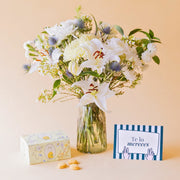 Ramo de flores blancas de Colvin en jarrón de cristal con lirios, crisantemos, eryngium azul y wax flower, junto a caja de galletas Loison de limón y tarjeta "Te lo mereces"