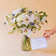 Ramo de flores blancas Colvin con lirio, crisantemo y eryngium en jarrón Gala, con mano sosteniendo tarjeta "Que tengas un Bonito día"