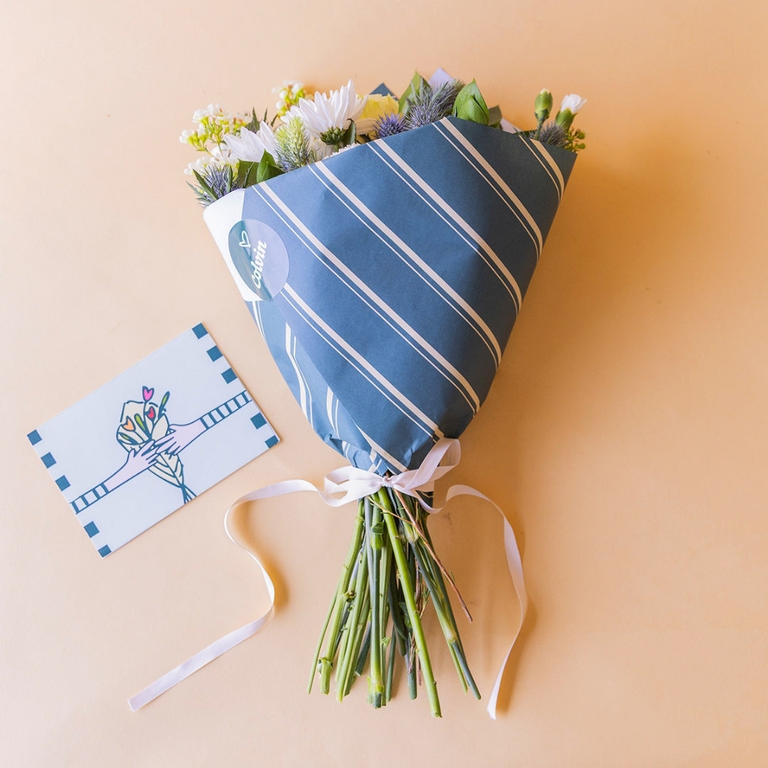 Ramo de flores blancas Colvin envuelto en papel de líneas azul con lazo blanco, junto a tarjeta Más que Flores sobre fondo crema
