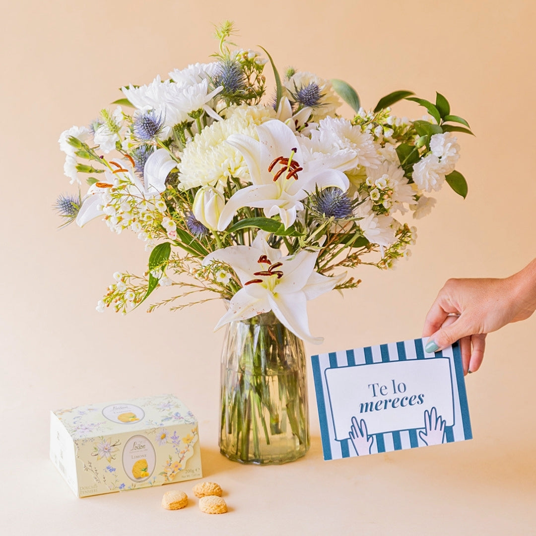 Mano sosteniendo tarjeta "Te lo mereces" junto a ramo de flores blancas de Colvin en jarrón de cristal con galletas Loison de limón