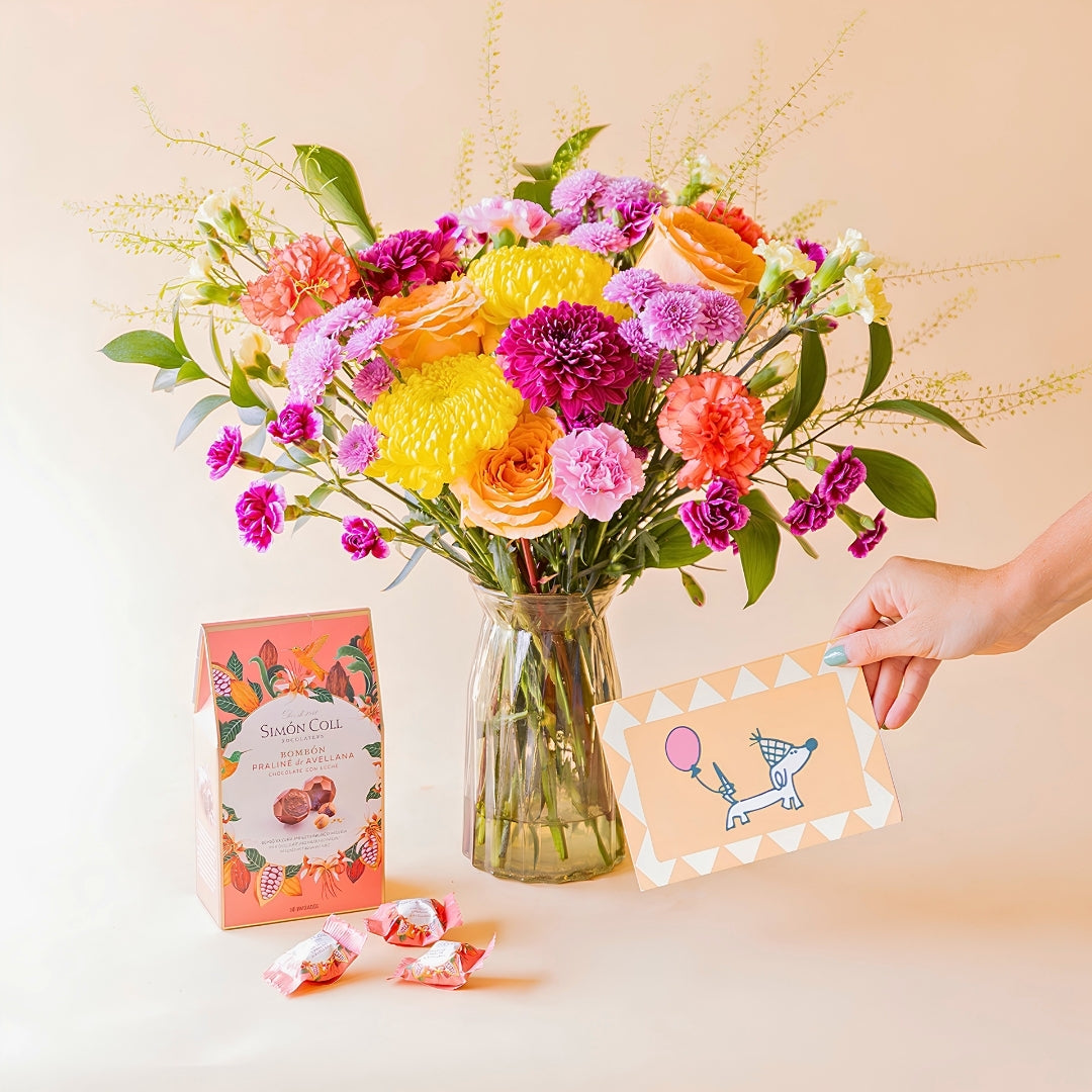 Mano sosteniendo tarjeta de cumpleaños con perrito junto a ramo de flores coloridas de Colvin en jarrón y bombones Simón Coll