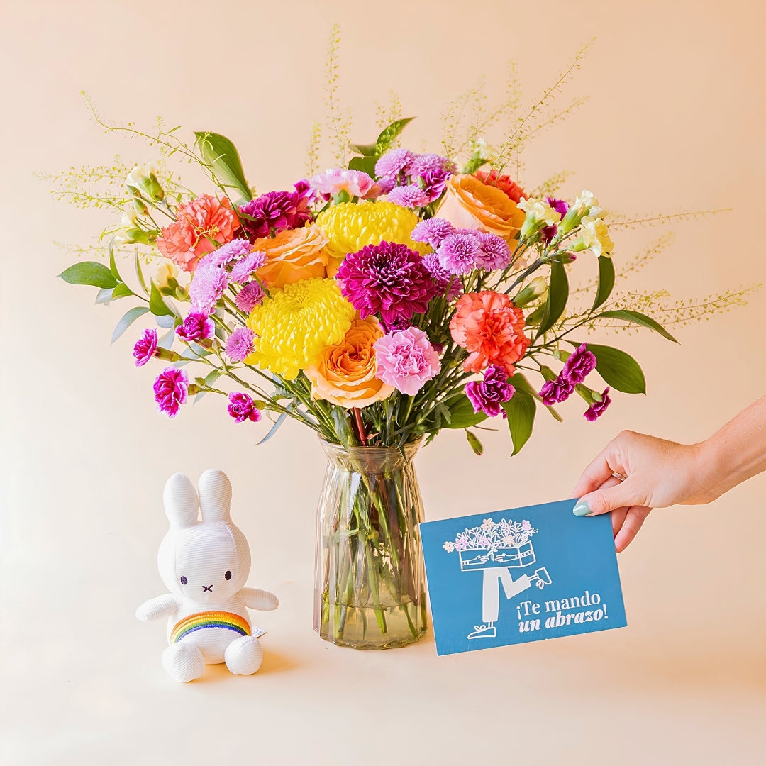 Mano sosteniendo tarjeta azul "¡Te mando un abrazo!" junto a ramo de flores coloridas de Colvin en jarrón de cristal con peluche Miffy arcoíris