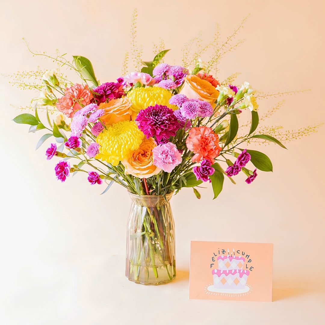 Ramo de flores El Colorido de Colvin en jarrón de cristal junto a tarjeta de cumpleaños, con rosas, crisantemos y flores en tonos vibrantes