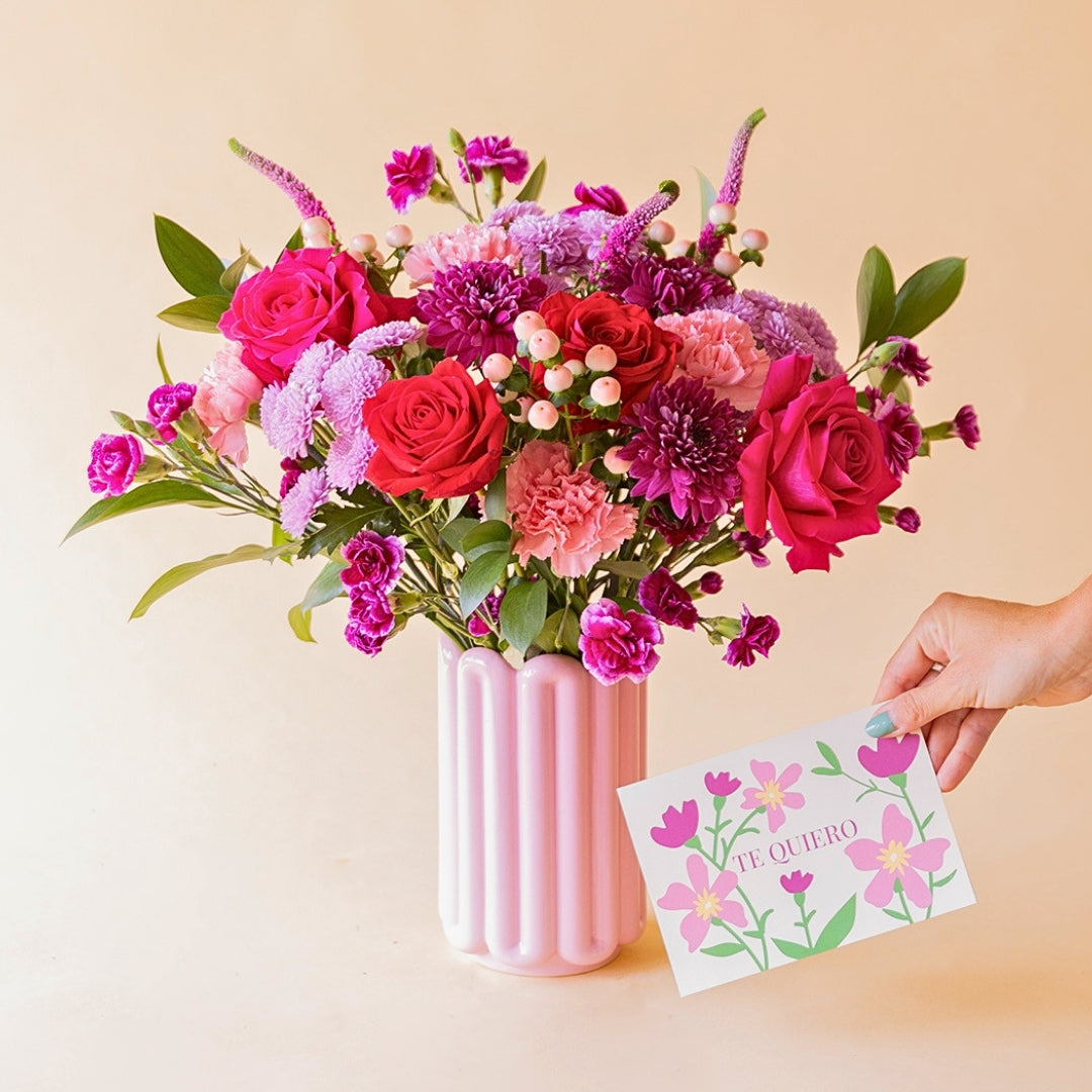 Ramo El Hechizo de Colvin en jarrón Lola rosa palo con mano sosteniendo tarjeta floral "Te Quiero" con ilustración de flores rosas y lilas