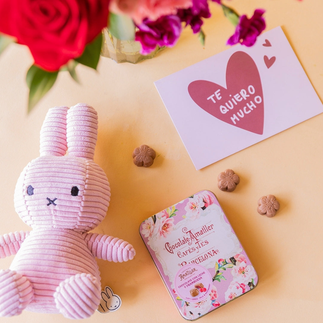 Detalle de peluche Miffy rosa de corduroy, lata de bombones Amatller chocolate con leche y frambuesa y tarjeta "Te quiero mucho" junto a ramo de flores rojas y fucsia de Colvin desenfocado