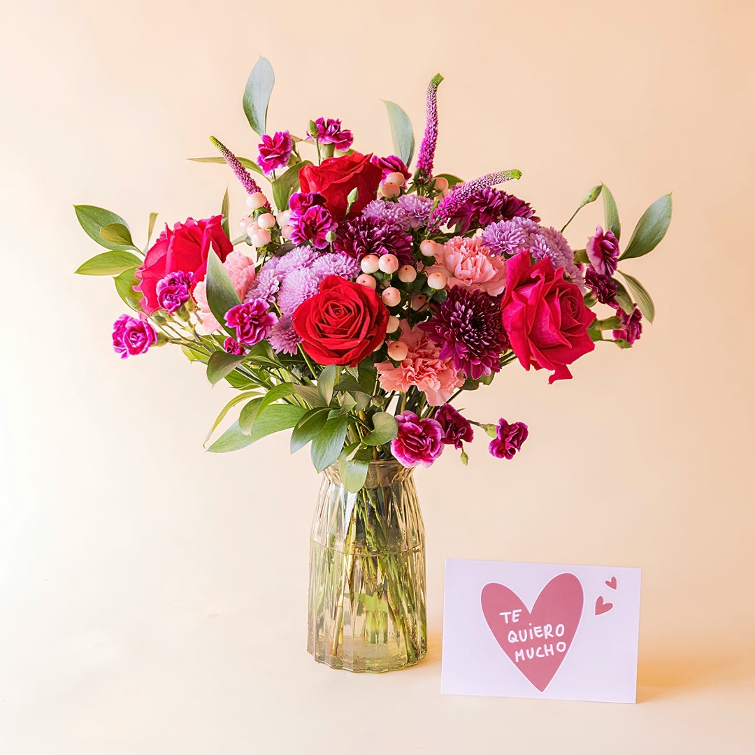 Ramo de flores rojas de Colvin en jarrón de cristal con rosas rojas, crisantemos lilas y claveles fucsia junto a tarjeta "Te quiero mucho"
