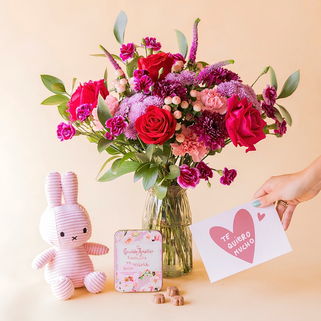 Mano sosteniendo tarjeta "Te quiero mucho" junto a ramo de flores rojas y fucsia de Colvin en jarrón de cristal con peluche Miffy rosa y lata de bombones Amatller frambuesa
