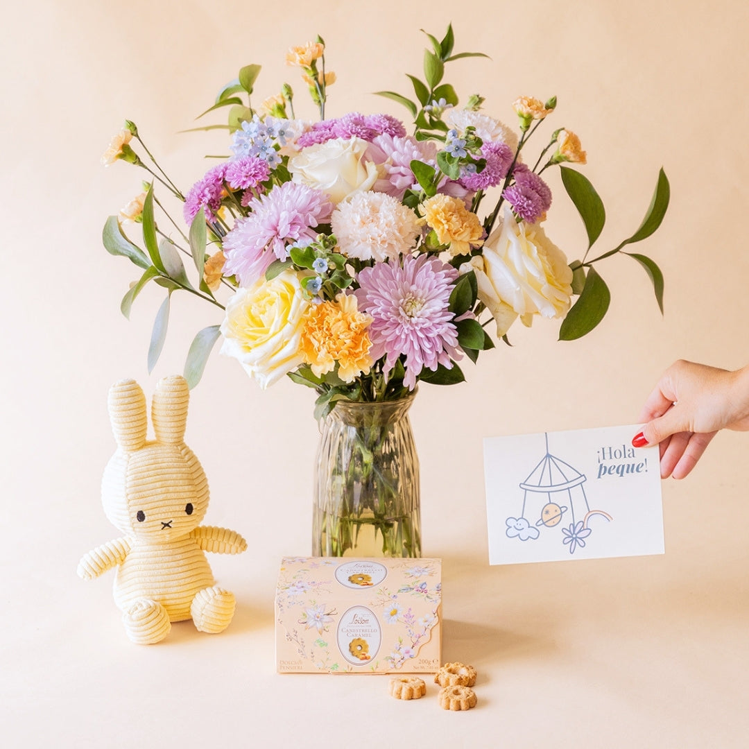 Mano sosteniendo tarjeta "¡Hola peque!" junto a ramo de flores pastel de Colvin en jarrón de cristal con peluche Miffy amarillo y galletas Loison Estrello Caramel