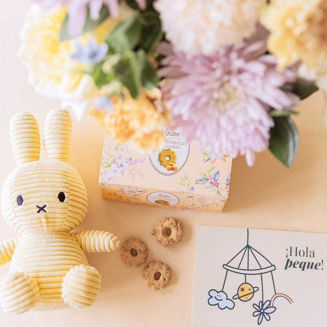 Detalle de peluche Miffy amarillo de corduroy, galletas Loison Estrello Caramel y tarjeta "¡Hola peque!" junto a ramo de flores pastel de Colvin desenfocado