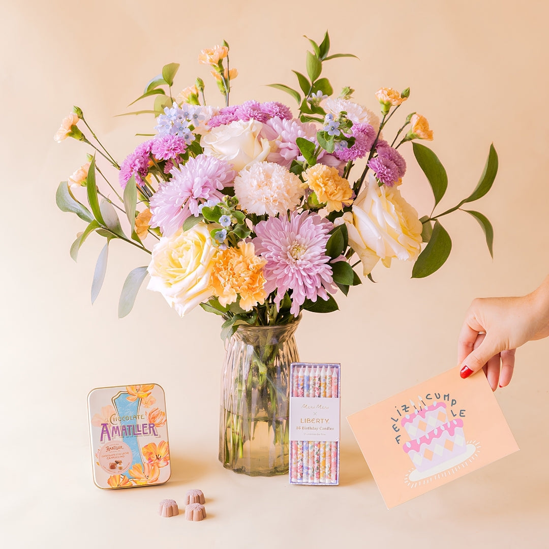 Mano sosteniendo tarjeta "Feliz Cumple" junto a ramo de flores pastel de Colvin en jarrón de cristal con lata de bombones Amatller y velas Liberty Meri Meri
