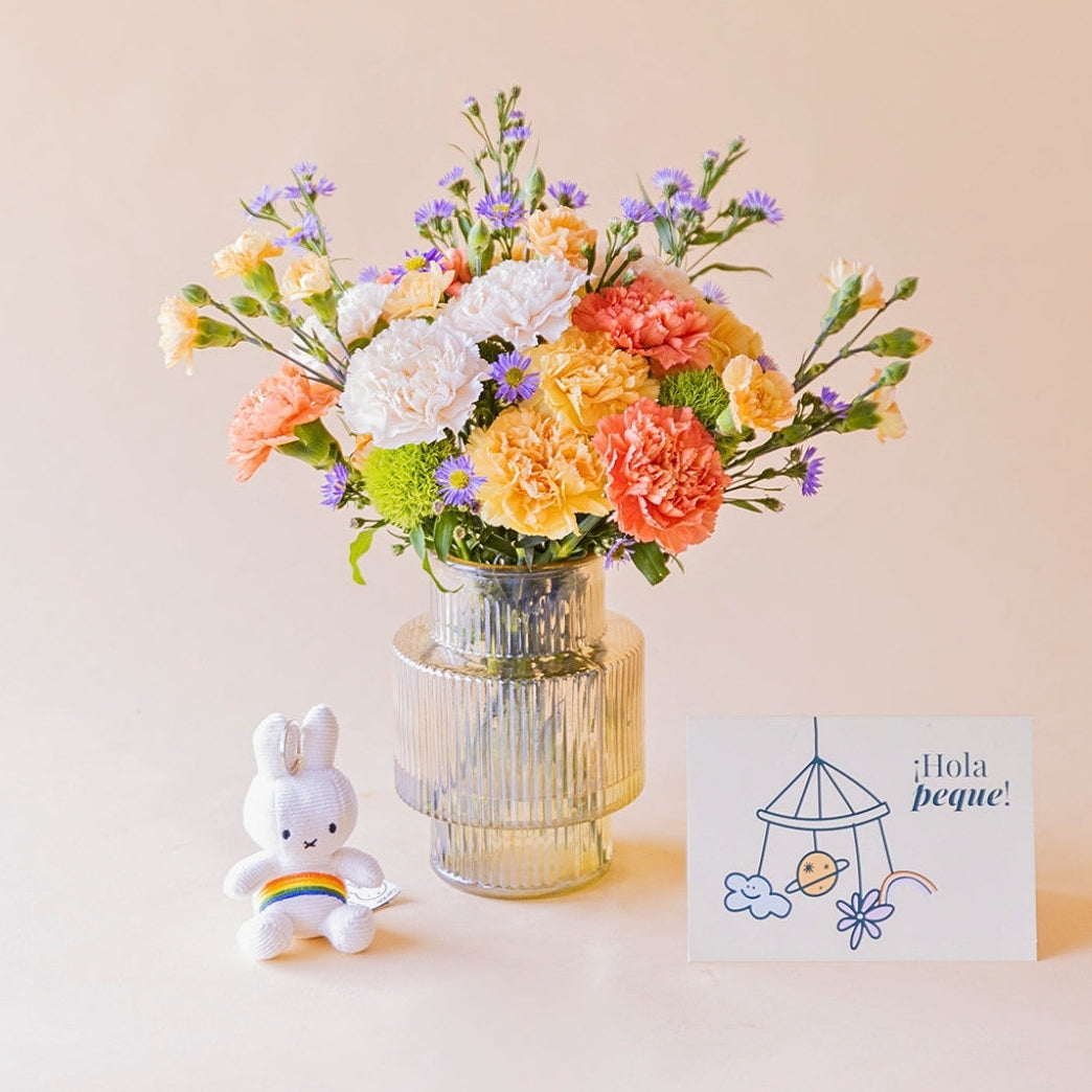 Ramo de claveles naranja, melocotón, blanco y solidago amarillo de Colvin con aster lila en jarrón acanalado transparente, con llavero peluche Miffy arcoíris y tarjeta ¡Hola peque! ilustrada, sobre fondo melocotón