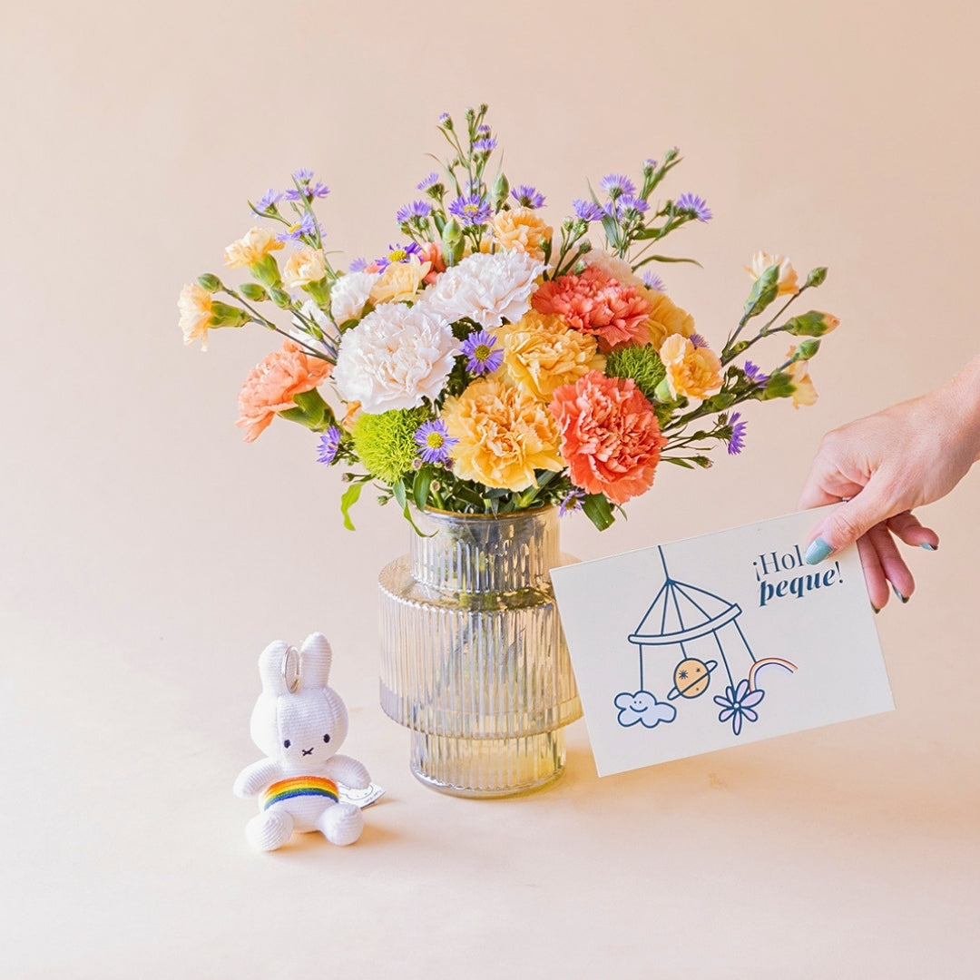 Ramo de claveles naranja, melocotón, blanco y aster lila de Colvin en jarrón acanalado transparente, con llavero peluche Miffy arcoíris y mano sosteniendo tarjeta ¡Hola peque!, sobre fondo melocotón