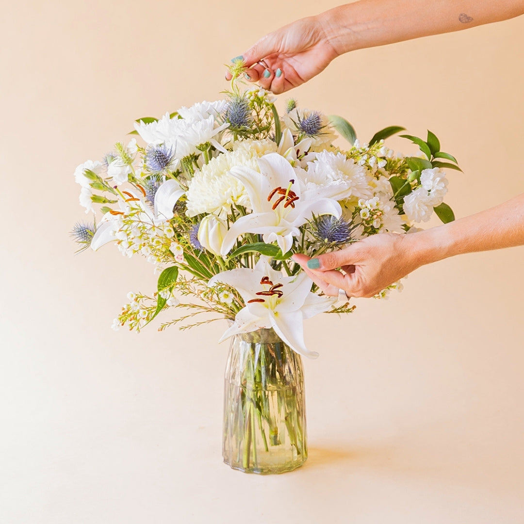 Manos colocando ramo de flores blancas con lirio oriental, crisantemo, eryngium y wax flower en jarrón de cristal