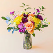 Ramo de flores frescas El Colorido de Colvin, un diseño vibrante y lleno de energía con rosas naranjas, crisantemos amarillos brillantes y flores en tonos púrpura y fucsia, presentado en un jarrón de cristal estriado sobre un fondo crema.