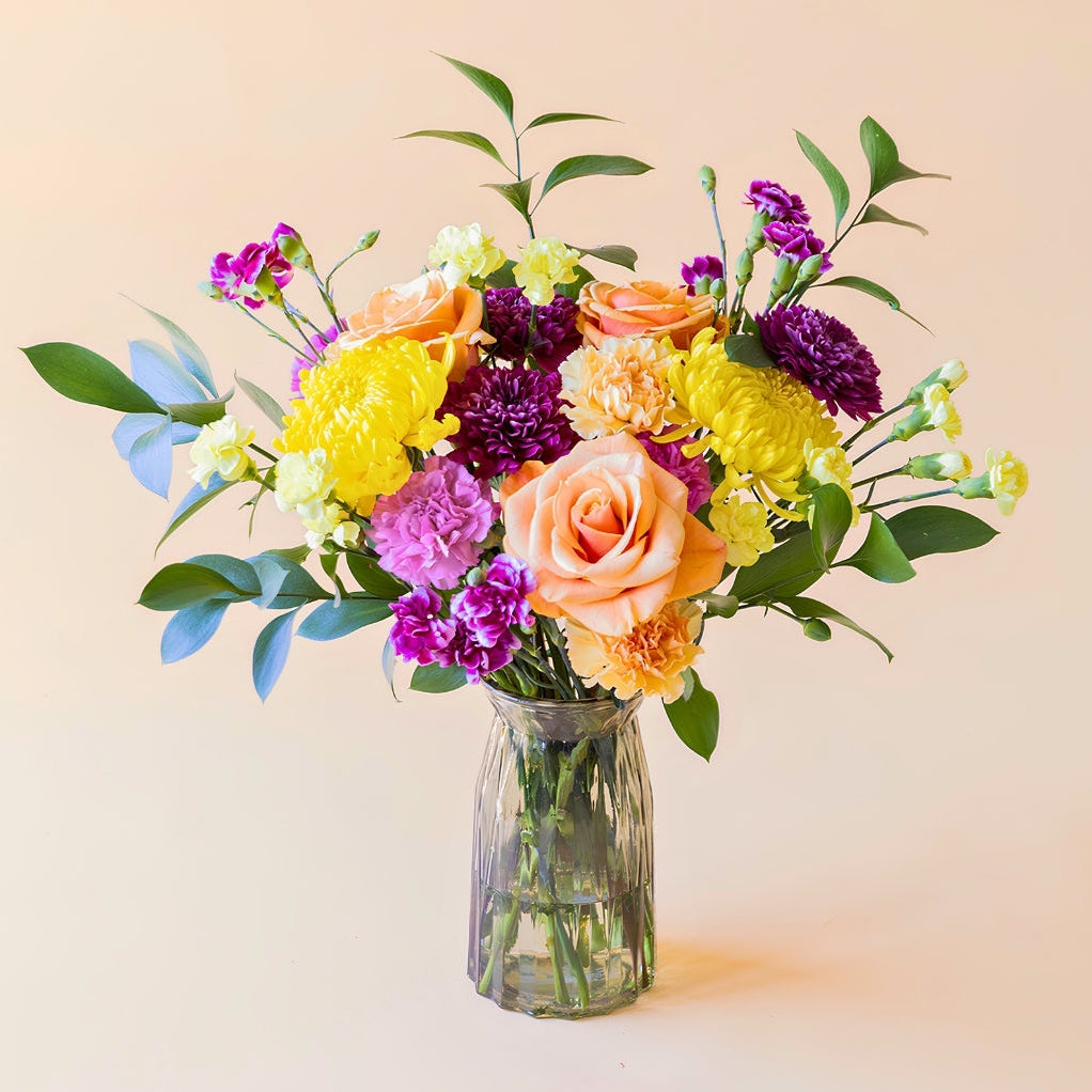Ramo de flores frescas El Colorido de Colvin, un diseño vibrante y lleno de energía con rosas naranjas, crisantemos amarillos brillantes y flores en tonos púrpura y fucsia, presentado en un jarrón de cristal estriado sobre un fondo crema.