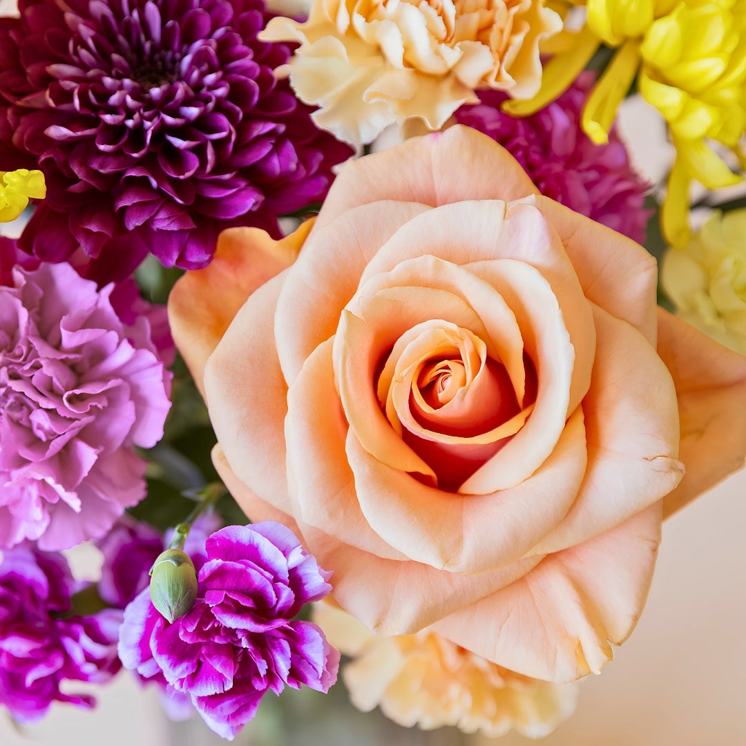 Detalle del ramo de flores frescas El Colorido de Colvin, un diseño vibrante y lleno de energía con rosas naranjas, crisantemos amarillos brillantes y flores en tonos púrpura y fucsia.