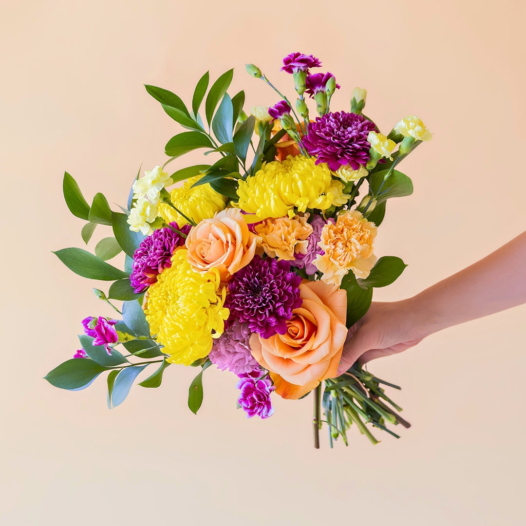 Mano sosteniendo el ramo de flores frescas El Colorido de Colvin, un diseño vibrante y lleno de energía con rosas naranjas, crisantemos amarillos brillantes y flores en tonos púrpura y fucsia, sobre un fondo crema.