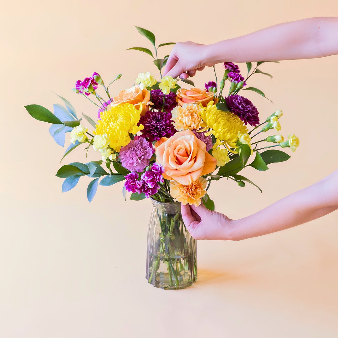 Manos tocando el ramo de flores frescas El Colorido de Colvin, un diseño vibrante y lleno de energía con rosas naranjas, crisantemos amarillos brillantes y flores en tonos púrpura y fucsia, presentado en un jarrón de cristal estriado sobre un fondo crema.