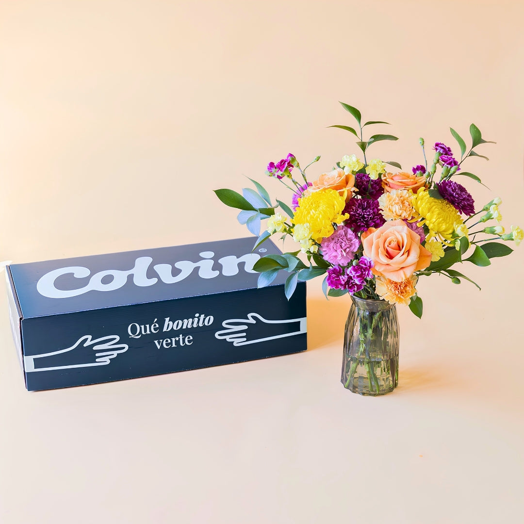Caja de Colvin verde oscuro con un mensaje "Qué bonito verte", ramo de flores frescas El Colorido, un diseño vibrante y lleno de energía con rosas naranjas, crisantemos amarillos brillantes y flores en tonos púrpura y fucsia, presentado en un jarrón de cristal estriado sobre un fondo crema.
