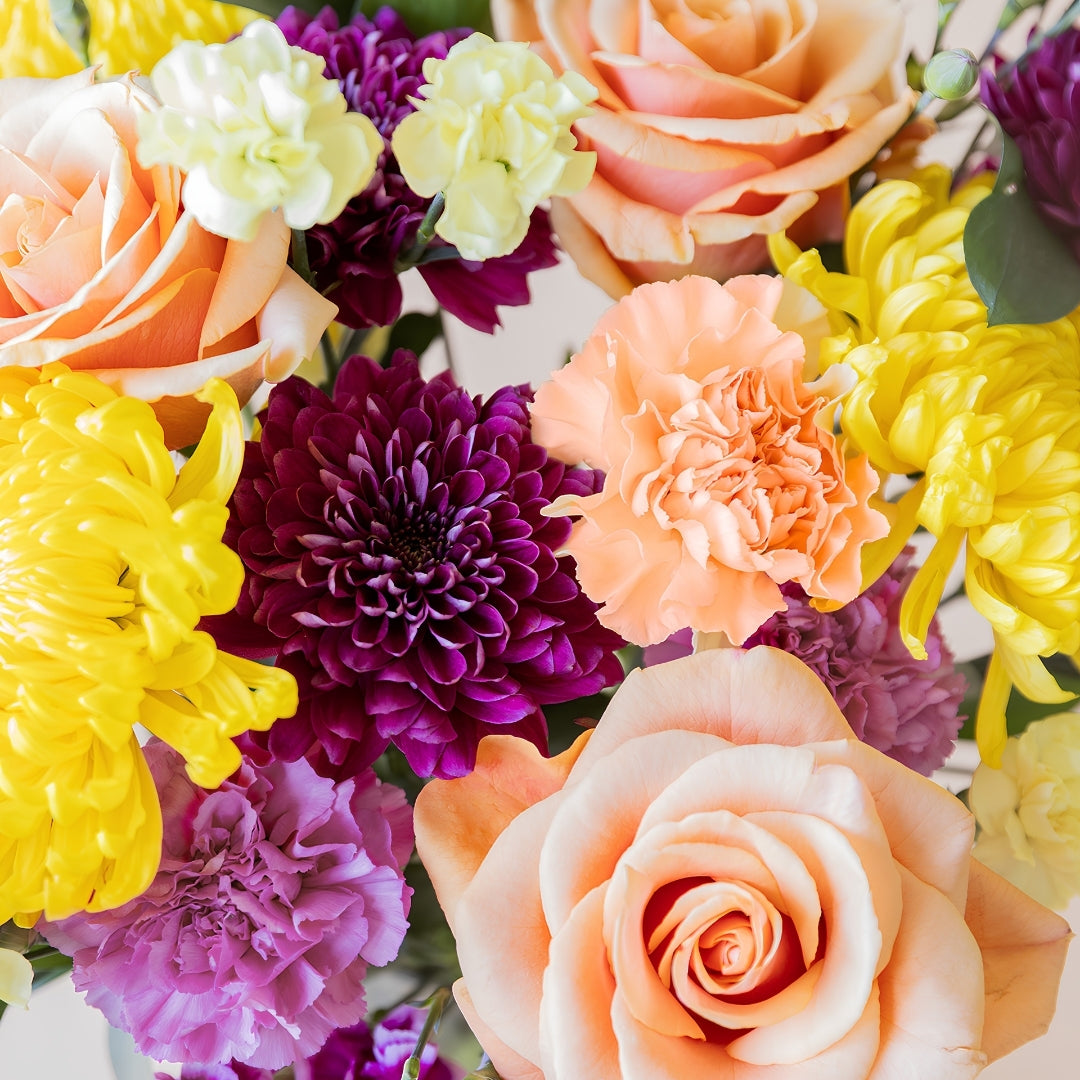Detalle del ramo de flores frescas El Colorido de Colvin, un diseño vibrante y lleno de energía con rosas naranjas, crisantemos amarillos brillantes y flores en tonos púrpura y fucsia.