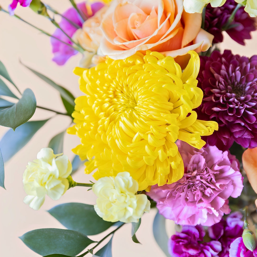 Detalle del ramo de flores frescas El Colorido de Colvin, un diseño vibrante y lleno de energía con rosas naranjas, crisantemos amarillos brillantes y flores en tonos púrpura y fucsia.