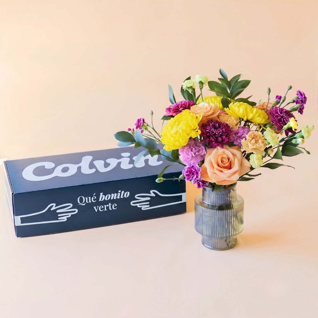 Caja de Colvin verde oscuro con un mensaje "Qué bonito verte", ramo de flores frescas El Colorido, un diseño vibrante y lleno de energía con rosas naranjas, crisantemos amarillos brillantes y flores en tonos púrpura y fucsia, presentado en un jarrón de cristal estriado sobre un fondo crema.