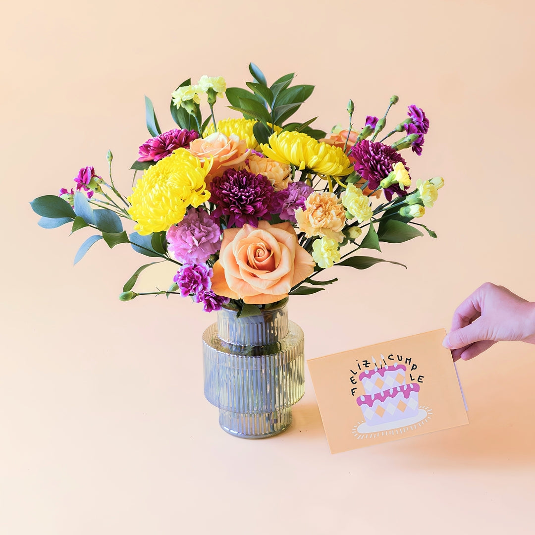 Ramo de flores frescas El Colorido de Colvin en un jarrón de cristal gris estriado de diseño geométrico, junto a una mano que sostiene una tarjeta de felicitación de cumpleaños con la frase "Feliz Cumple" y una tarta ilustrada, sobre fondo beige.