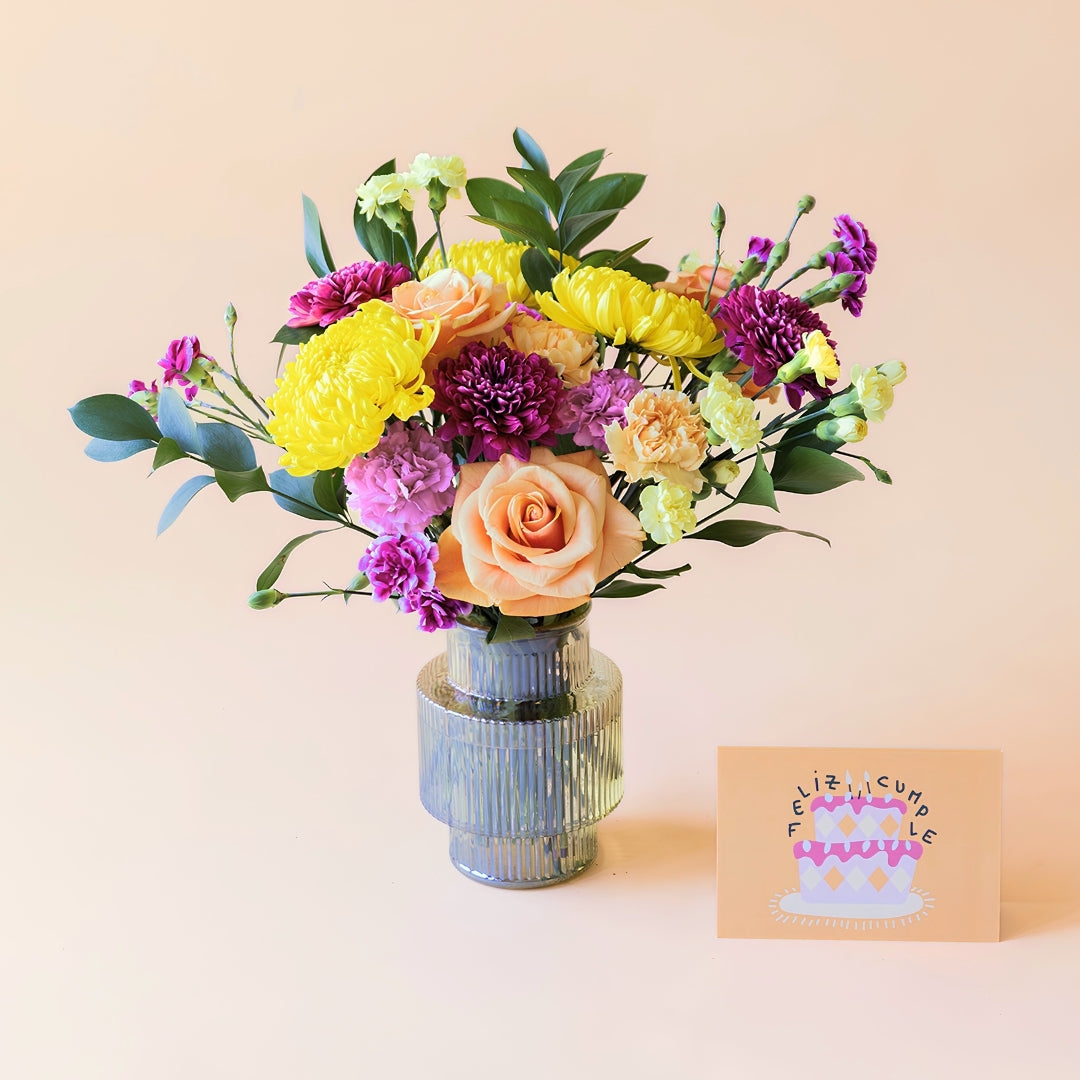Ramo de flores frescas El Colorido de Colvin en un jarrón de cristal gris estriado de diseño geométrico, una tarjeta de felicitación de cumpleaños con la frase "Feliz Cumple" y una tarta ilustrada, sobre fondo beige.