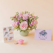Arreglo de flores frescas en tonos rosas Dulce con base estriada verde menta, junto a una hoja de pegatinas ilustradas con frases, un pequeño llavero de peluche rosa con textura de pana y carita sonriente, y una tarjeta de agradecimiento violeta con la palabra "Gracias" sobre un fondo liso de color crema.
