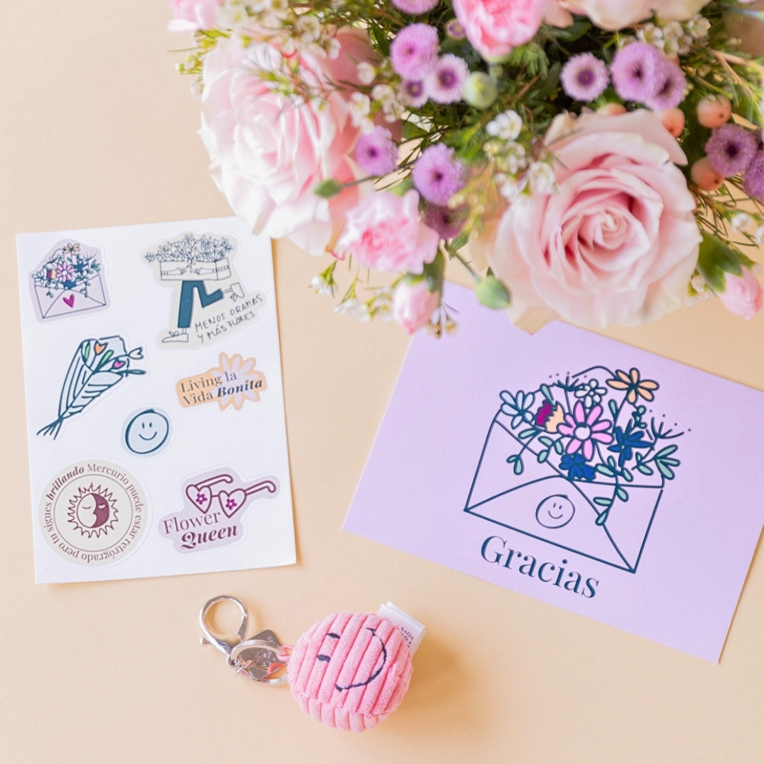 Detalle del arreglo de flores frescas en tonos rosas Dulce, junto a una hoja de pegatinas ilustradas con frases, un pequeño llavero de peluche rosa con textura de pana y carita sonriente, y una tarjeta de agradecimiento violeta con la palabra "Gracias" sobre un fondo liso de color crema. 