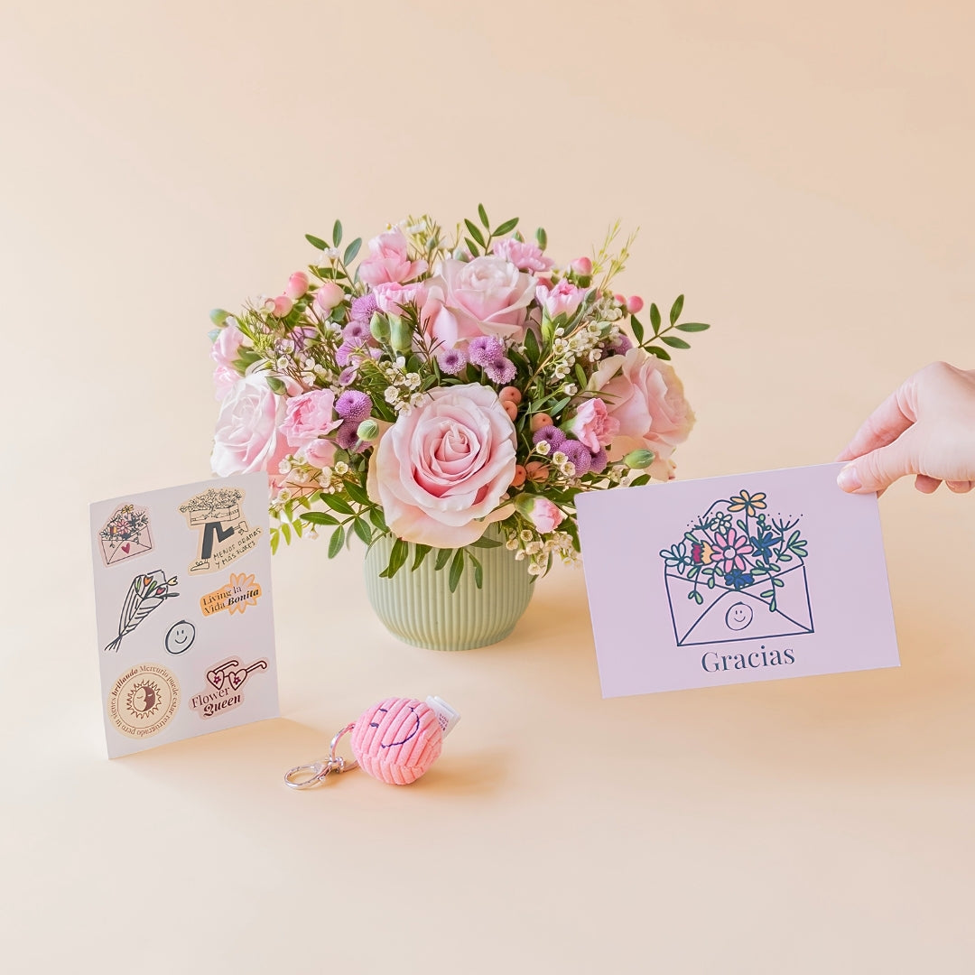Arreglo de flores frescas en tonos rosas Dulce con base estriada verde menta, junto a una hoja de pegatinas ilustradas con frases, un pequeño llavero de peluche rosa con textura de pana y carita sonriente, y una mano sosteniendo una tarjeta de agradecimiento violeta con la palabra "Gracias" sobre un fondo liso de color crema.