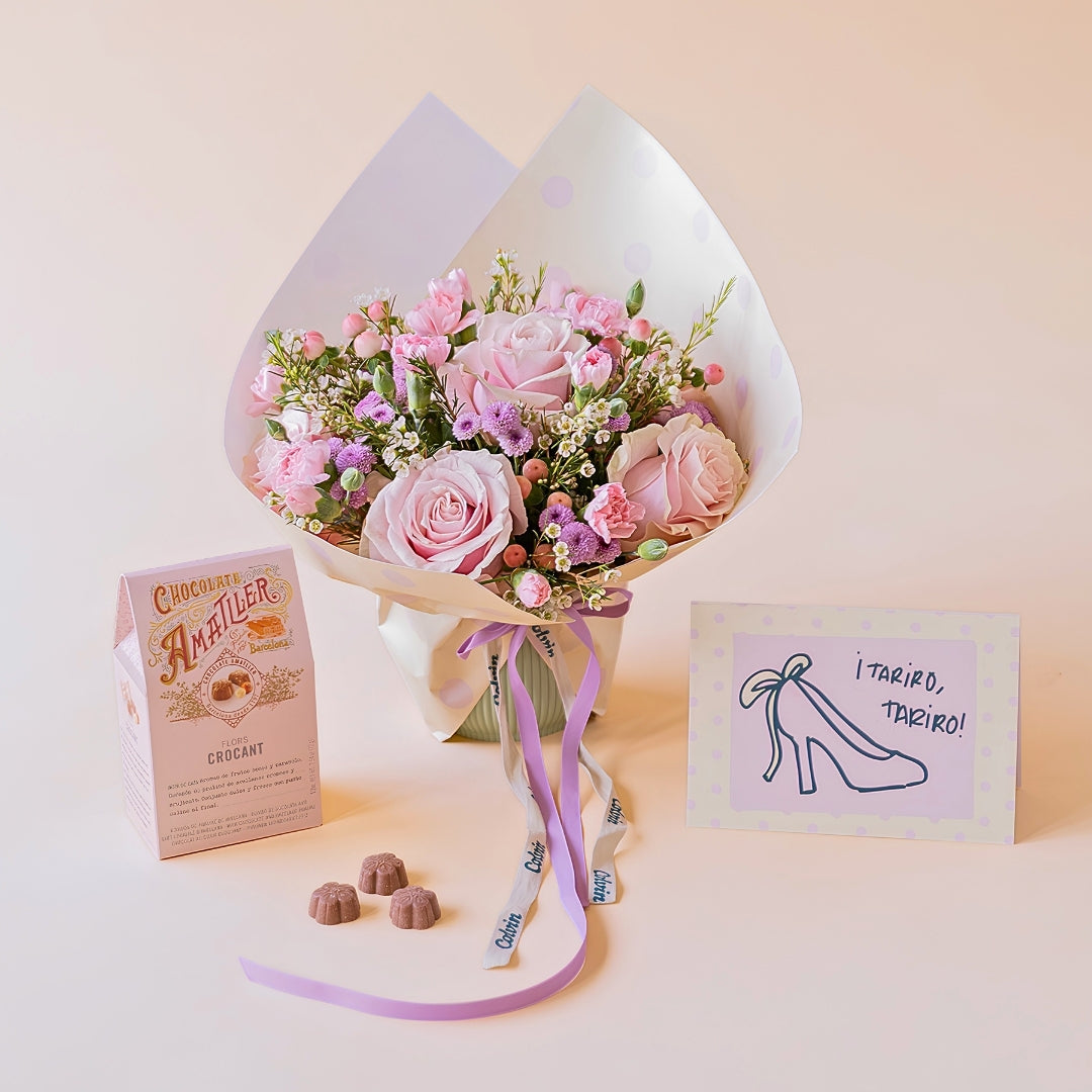 Arreglo floral rosas Dulce con envoltorio de papel de lunares y lazo violeta, junto a una caja de bombones de chocolate Amatller y una tarjeta de felicitación para novias con la ilustración de un zapato de tacón y la frase ¡Tariro, tariro! sobre un fondo color crema.