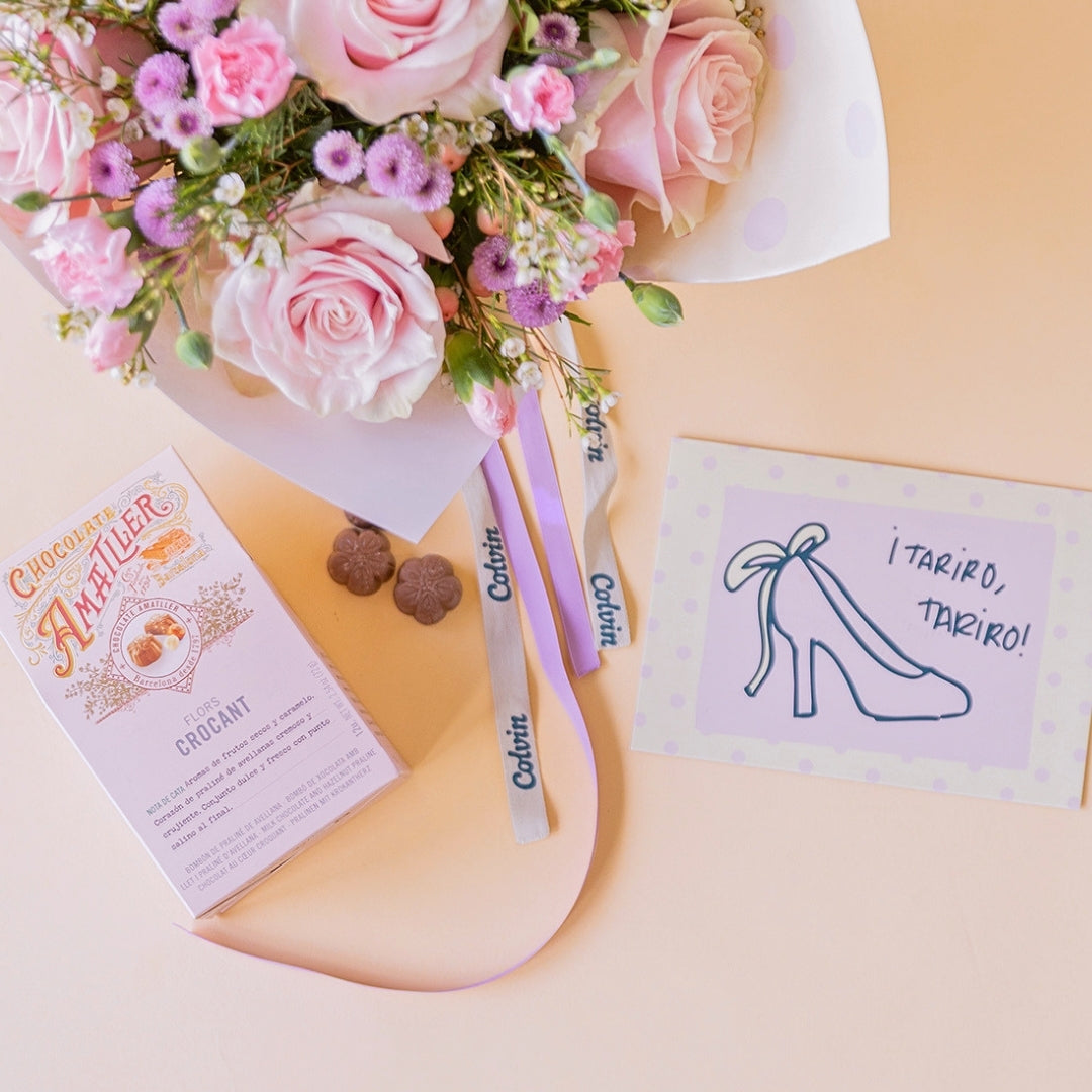 Detalle del arreglo floral rosas Dulce con envoltorio de papel de lunares y lazo violeta, junto a una caja de bombones de chocolate Amatller y una tarjeta de felicitación para novias con la ilustración de un zapato de tacón y la frase ¡Tariro, tariro! sobre un fondo color crema.