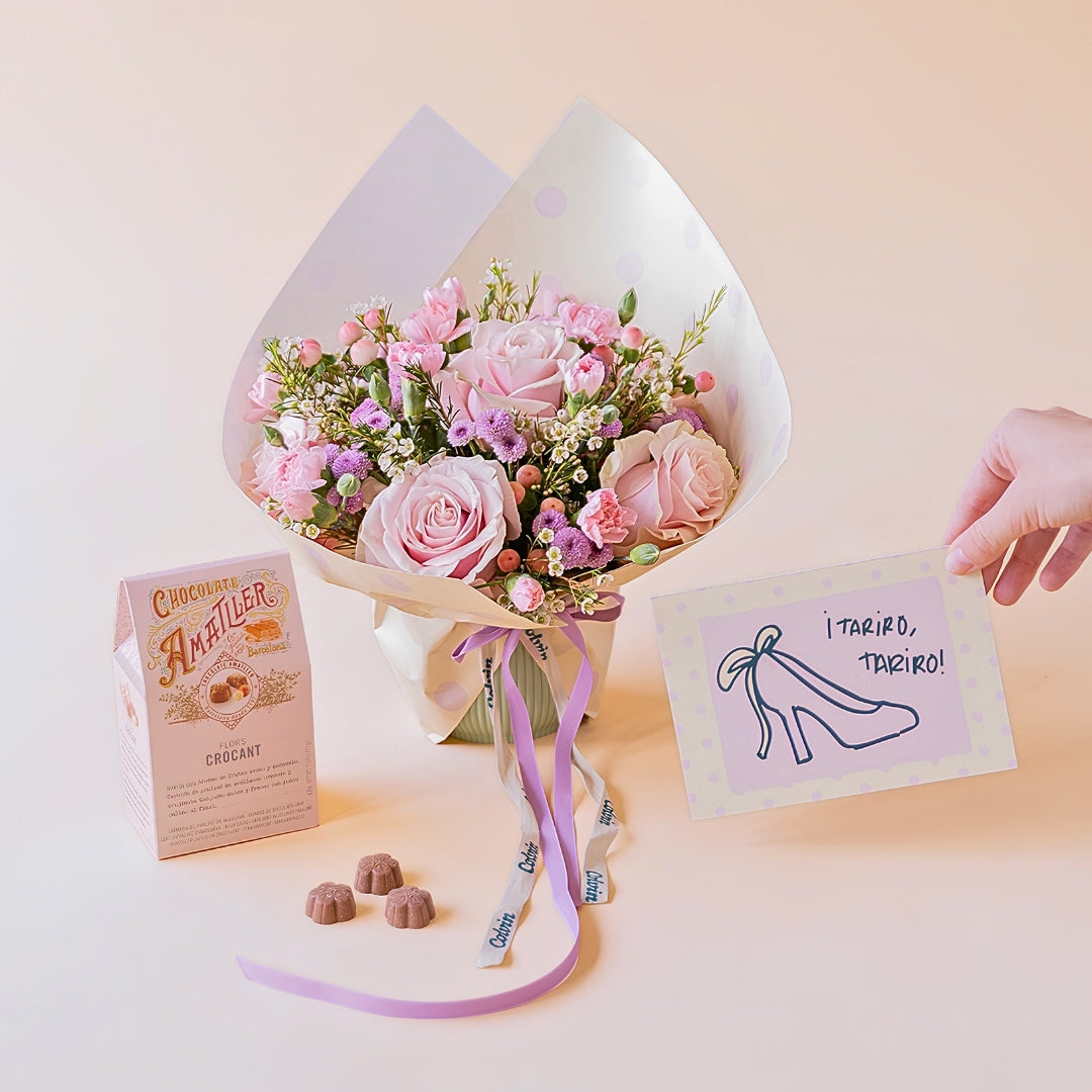 Arreglo floral rosas Dulce con envoltorio de papel de lunares y lazo violeta, junto a una caja de bombones de chocolate Amatller y una mano sosteniendo una tarjeta de felicitación para novias con la ilustración de un zapato de tacón y la frase ¡Tariro, tariro! sobre un fondo color crema.