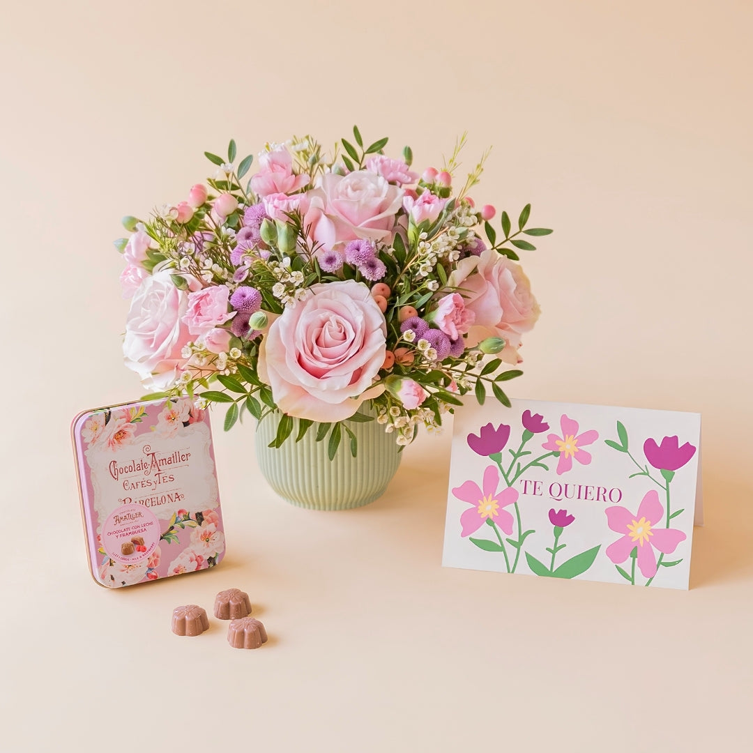 Arreglo de flores frescas en tonos rosas Dulce base estriada verde menta, junto a una lata vintage de bombones de chocolate Amatller Barcelona y una tarjeta regalo ilustrada con la frase "TE QUIERO" sobre un fondo liso de color crema.