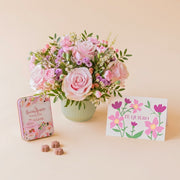 Arreglo de flores frescas en tonos rosas Dulce base estriada verde menta, junto a una lata vintage de bombones de chocolate Amatller Barcelona y una tarjeta regalo ilustrada con la frase "TE QUIERO" sobre un fondo liso de color crema.